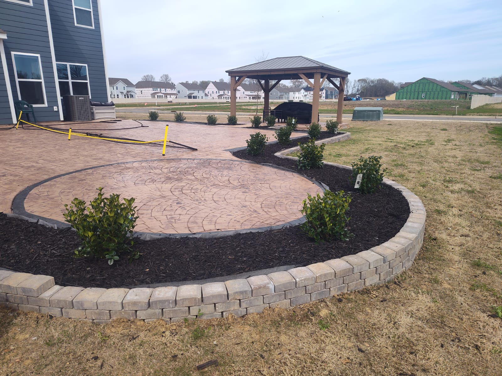 Circular stamped concrete patio with paver seat wall, mulched planting beds, and cedar pavilion