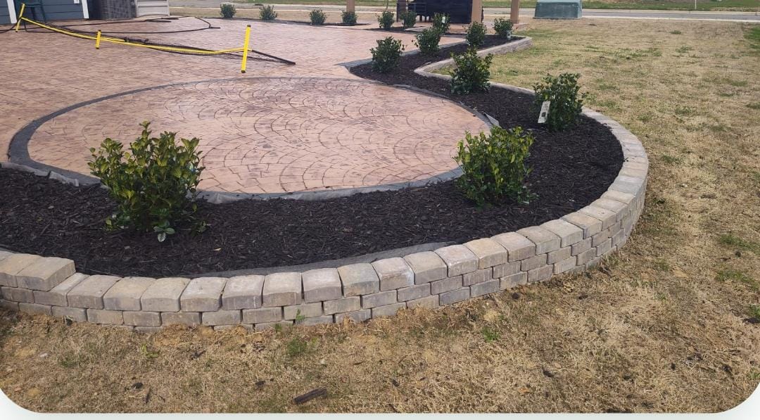 Circular stamped concrete patio bordered by a curved paver wall, fresh mulch beds, and new shrubs